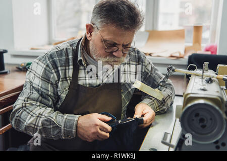 mature male tailor in eyeglasses and apron cutting leather by scissors at workshop Stock Photo