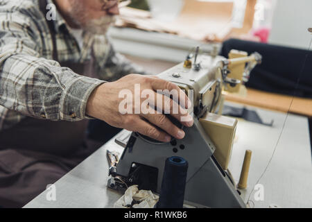 partial view of male tailor in apron and eyeglasses working on sewing machine at studio Stock Photo