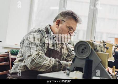 focused mature male tailor in apron and eyeglasses working on sewing machine at studio Stock Photo