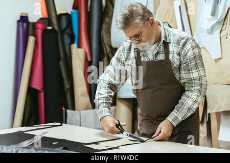 concentrated male handbag craftsman in apron and eyeglasses cutting leather by scissors at workshop Stock Photo