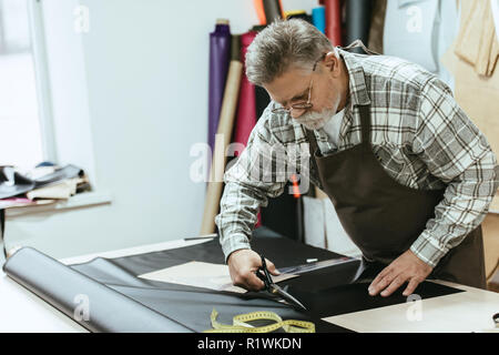 male craftsman in apron and eyeglasses cutting leather by scissors at workshop Stock Photo