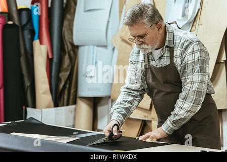 mature male handbag craftsman in apron and eyeglasses cutting leather by scissors at workshop Stock Photo