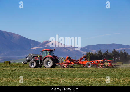 Red farm machinery working in a rural area of New Zealand cutting and raking crops to make hay Stock Photo