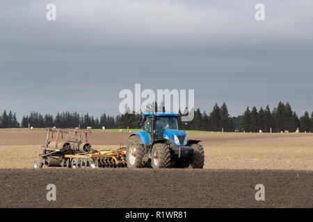 A blue tractor working with a plough in a farm field in spring to prepare it for planting a crop in Canterbury, New Zealand Stock Photo