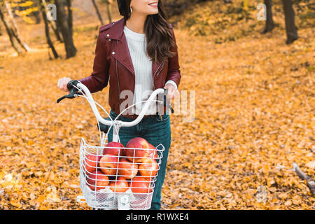 partial view of fashionable girl in leather jacket carrying bicycle with basket full of red apples in autumnal park Stock Photo