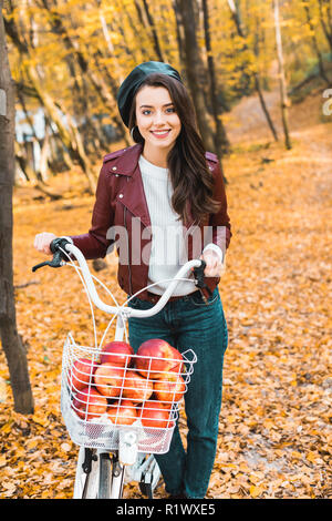 smiling stylish girl in leather jacket carrying bicycle with basket full of red apples in autumnal park Stock Photo