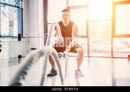 young sportsman working out with battle ropes at gym in sunlight Stock Photo
