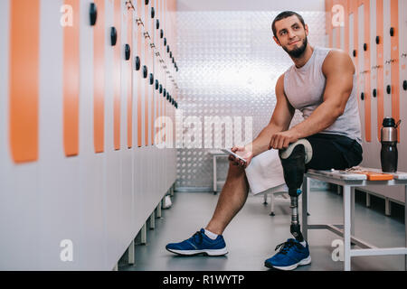 smiling young sportsman with artificial leg sitting on bench at gym changing room and using smartphone Stock Photo
