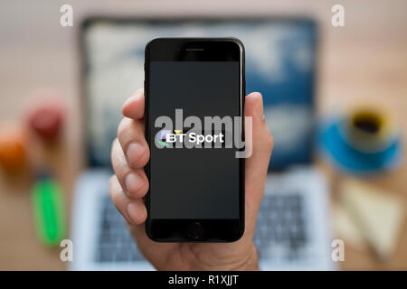 A man looks at his iPhone which displays the BT Sport logo, while sat at his computer desk (Editorial use only). Stock Photo