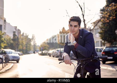 street (out of focus in background), one young man posing in foreground, looking sideways,  bicycle handlebar outdoors. 20-29 years old, wearing suit. Stock Photo