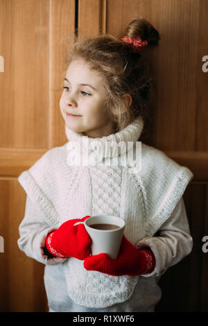 Charming seven-year-old girl drinks warm tea, standing next to the window. She is wearing a warm knitted sweater, her hands are in red knitted gloves. Stock Photo