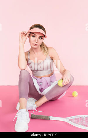 stylish sportswoman in visor hat posing with tennis racket and balls on pink Stock Photo