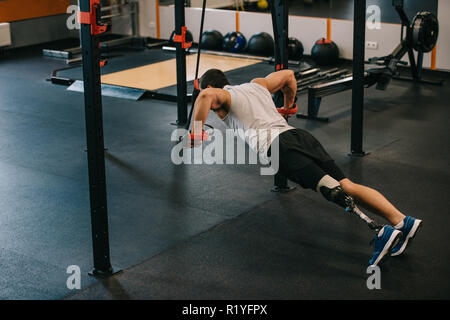 handsome young sportsman with artificial leg doing push ups with hanging rings of gymnastics ladder at gym Stock Photo