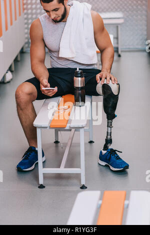 attractive young sportsman with artificial leg sitting on bench at gym changing room and using smartphone Stock Photo