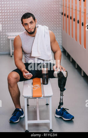 handsome young sportsman with artificial leg sitting on bench at gym changing room and using smartphone Stock Photo