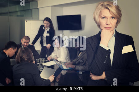 Unhappy mature business woman standing in meeting room on background with coworkers Stock Photo