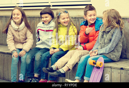 Group of cheerful  children sitting on bench and sharing secrets outdoors Stock Photo