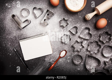 Cookie cutters shaped in word Love with flour lying on black background. Baking cookies concept. Cookery book on the table. Stock Photo