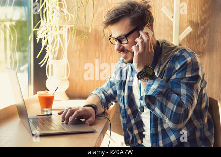Modern young man in coffee shop using laptop and chatting on smartphone sitting near window Stock Photo