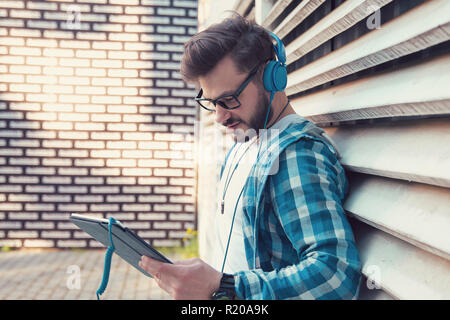 Cheerful casual man in denim shirt and headphones standing outdoors and surfing tablet happily Stock Photo
