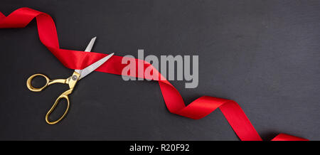 Grand opening. Top view of gold scissors cutting red silk ribbon against black background, banner, copy space. Stock Photo