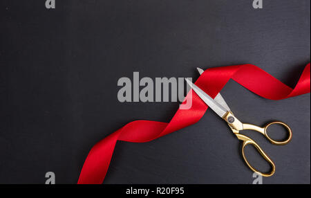 Grand opening. Top view of gold scissors cutting red silk ribbon against black background, copy space Stock Photo
