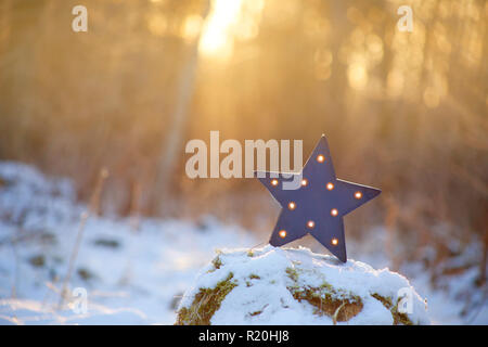 Focus on decorative modern blue star shape lamp with lots of burning small light bulbs on, outdoors on snowy ground, with nice back light from sun set Stock Photo