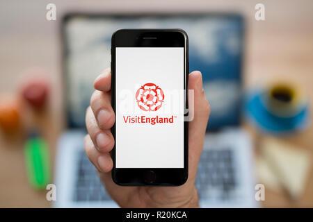 A man looks at his iPhone which displays the Visit England logo, while sat at his computer desk (Editorial use only). Stock Photo