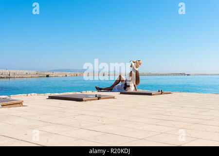 Happy tourist woman relaxing at the Venetian port of Heraklion. Lovely stylish girl sitting on harbor dock enjoying vacation travel to Crete. Girl on  Stock Photo