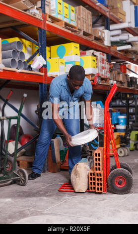 Concentrated African American man loading building materials on hand trolley cart in shop Stock Photo
