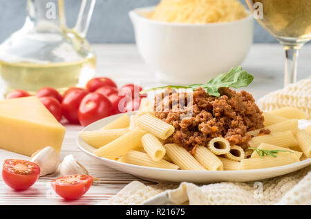 Italian pasta bolognese penne rigatone minced meat in tomato sauce and parmesan cheese. Still life on white wooden table served with cherry tomatoes, carafe of olive oil and glass of white wine Stock Photo