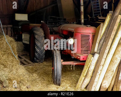 Dirty old abandoned vintage red David Brown 25T tractor in a barn surrounded by hay and straw, with wood and fence posts piled up in front of it. UK