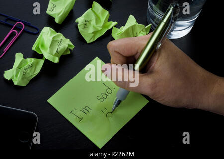 A hand writing on a green sticky note the phrase 'I'm sad.' A photo about depression, sadness and loneliness. Crumpled green sticky notes scattered. Stock Photo