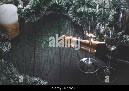 A bottle of champagne and two glasses stand on a dark gray wooden table surrounded by Christmas decorations and a white-gold candle stands next to it. Stock Photo