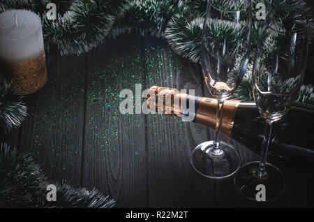 A bottle of champagne and two glasses stand on a dark gray wooden table surrounded by Christmas decorations and a white-gold candle stands next to it. Stock Photo