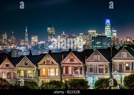 Backed by the night skyline of the city of San Francisco, California, the Victorian era houses near Alamo Square Park, are painted in colors to accent Stock Photo