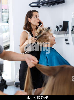 Emotional woman hairstylist having phone conversation while doing hairdress for elderly female client in hair salon Stock Photo