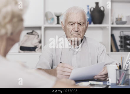 Upset senior man sitting at home table discussing with wife their bills Stock Photo