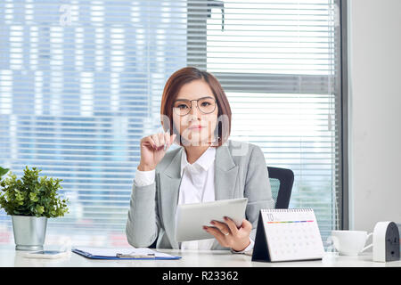 Portrait of beautiful young Asian business woman sitting and using a tablet behind the table in workplace Stock Photo