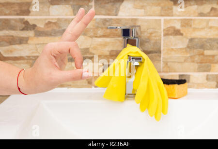 Person making okay gesture with fingers in front of white porcelain sink with yellow goves hanging on iron tap and sponge near Stock Photo