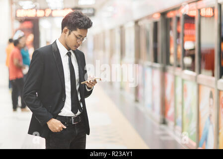 Young hipster asian businessman using smartphone while waiting for a train in subway. Concept of wireless technology, mobile internet and online busin Stock Photo