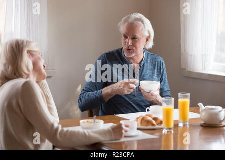 Happy senior couple enjoy morning at home eating healthy breakfast, having coffee, smiling aged husband and wife talk at kitchen table spending weeken Stock Photo