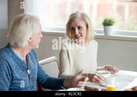 Happy elderly couple talk in kitchen having healthy breakfast at home, senior husband and wife enjoy delicious food spending time together, aged man a Stock Photo