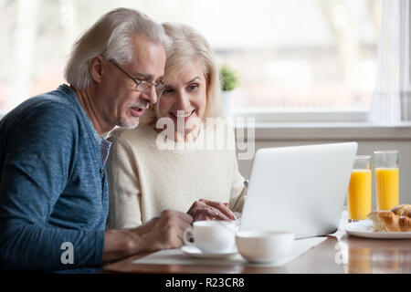 Aged couple busy look at laptop screen while having delicious breakfast at home kitchen, excited senior man and woman use computer during morning rout Stock Photo