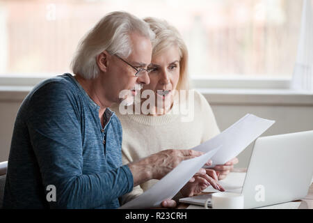 Concerned senior husband and wife hold documents using laptop checking information online, worried aged couple shocked by prices or tax growth reading Stock Photo