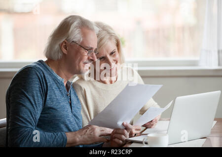 Happy aged husband and wife hold papers using laptop for online banking, satisfied senior couple smiling checking utility bills or insurance at comput Stock Photo