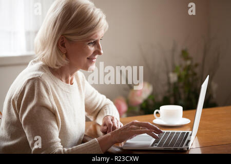 Excited aged woman using laptop, browsing internet at home, smart senior lady working at computer drinking coffee indoors, busy elderly female surfing Stock Photo