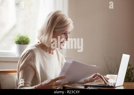 Senior woman holding papers busy at laptop managing house utility bills or finances, aged female using computer working with bank loan or mortgage doc Stock Photo