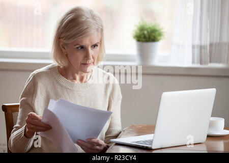 Serious aged woman holding documents, checking information at laptop online, concerned senior female managing bank insurance or loan papers, busy work Stock Photo