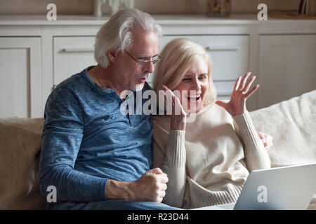 Excited aged husband and wife relax on couch at home feel euphoric winning online lottery at laptop, happy senior couple sit on sofa scream with happi Stock Photo